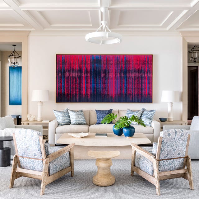 A-List 2024 - ELLE DECOR a living room with a off white sofa with a large blue and red abstract painting on the wall behind and wood and upholstered chairs