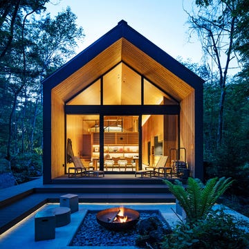 Modern cabin nestled in the woods at dusk, featuring a fire pit and wooden deck.