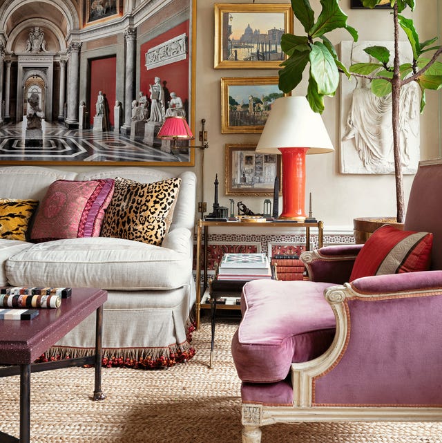 A-List 2024 - ELLE DECOR elegant living room with a plush linen sofa with fringe on the skirt and a pink velvet upholstered regency chair in the foreground and classical artwork in gold frames on the back wall and a side table with books and an modernistic orange ceramic base lamp and a jute rug on the floor
