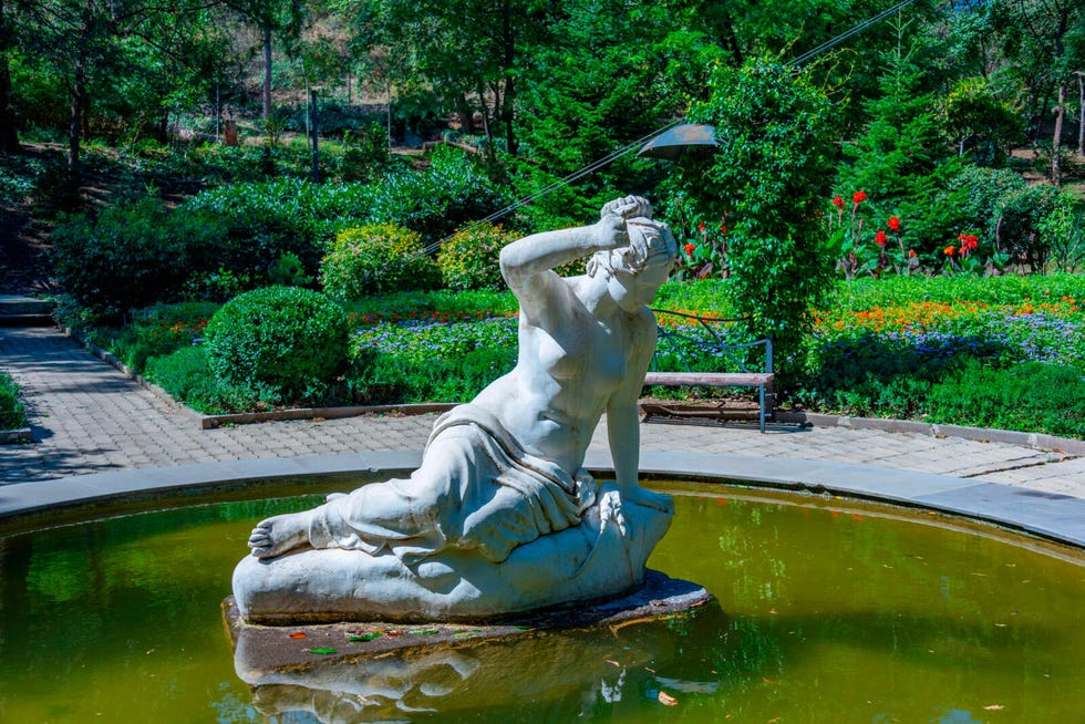 Sculpture of a reclining figure in a pond surrounded by greenery and flowers.