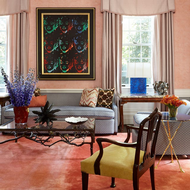 A-List 2024 - ELLE DECOR a pink painted living room with orange velvety rug a gray sofa with blue trim and matching chair and a dark wood armchair with yellow seat and floor to ceiling lighter pink curtains and a large dark framed artwork over the sofa and between the windows