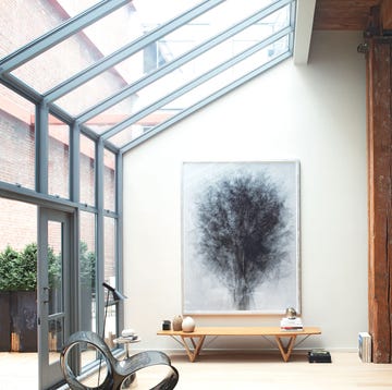 conservatory like area with wood bench with object on it and a glass wall and slanted roof adjoinging rough wood beams with furniture and loaded bookshelves