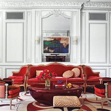 a living room with high ceilings and fancy moldings in white and a red velvet scroll sofa with end seats attached and a swirly circular white and red outline carpet and a leopard scroll leg modern stool and a shiny red lacquer bean shape table at center and a painting on the wall