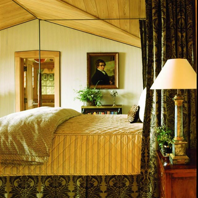 A-List 2024 - ELLE DECOR a colonial looking bedroom with a four poster bed with green patterened curtains and matching bed skirt and a yellow quilted blanket and a portrait on the far wall and a lamp
