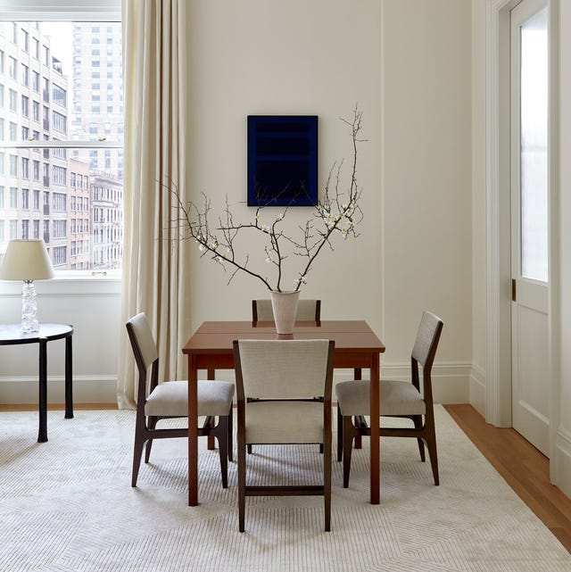 A-List 2024 - ELLE DECOR small wood table with a vase with branches on int and with linen and wood chairs and a off white rug in a off white room with a window and floor to ceiling curtains