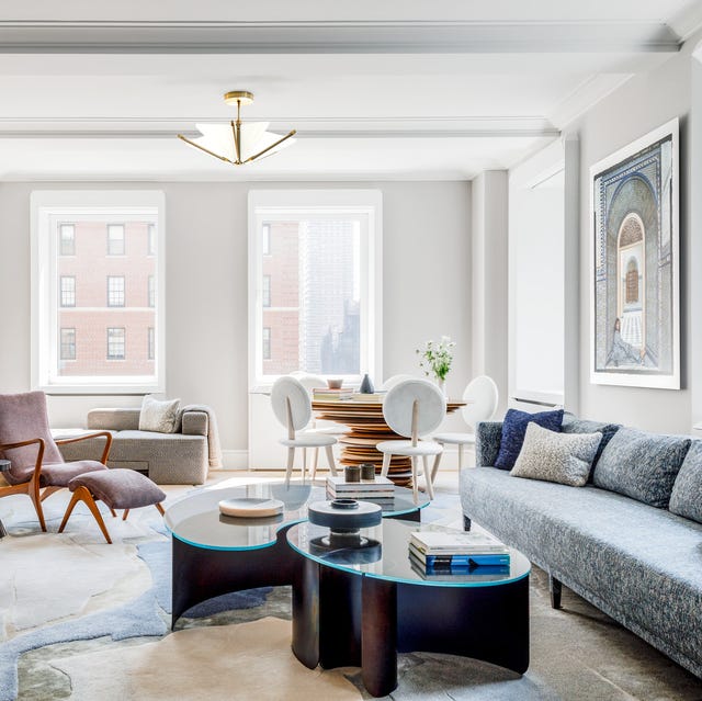 A-List 2024 - ELLE DECOR spacious airy living room with long blue upholstered sofa against the right wall and a cocktail table with interlocking sections topped with glass and a rug that looks like colored rock and chairs along the other side