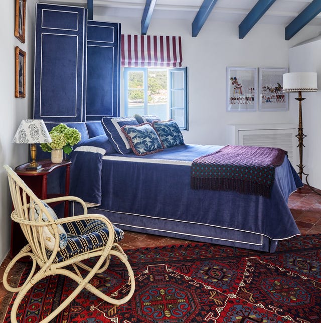 A-List 2024 - ELLE DECOR white room with blue ceiling beams and a bed set on an angle with a lush royal blue velvet cover with white trim and purple throw at the foot and matching blue folding screen with white trim behind the bed and a terracotta floor w ith a majestic blue and red patterned rug and a simple rattan chair in the foreground