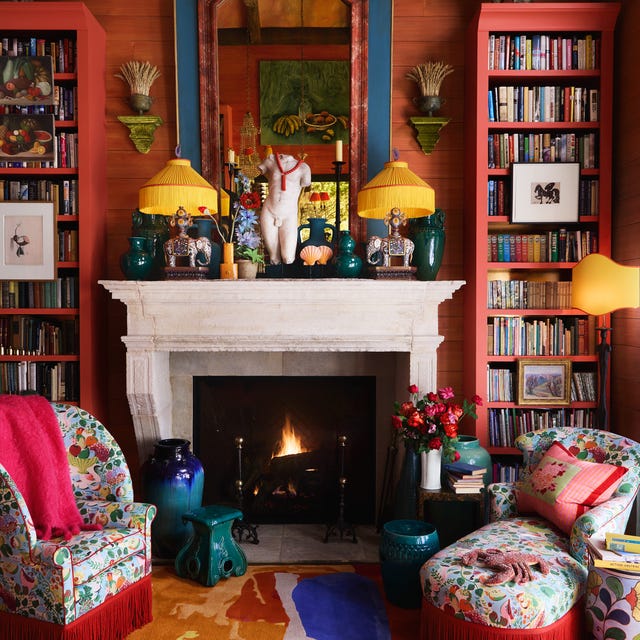 A-List 2024 - ELLE DECOR very cozy closeup of a sitting area with red plank walls and a white marble fireplace with a large blue and red mirror over it and jade colored objects and elephant lamps with matching yellow fringe shades and in front of the fire is a small colorful chair and matching chaise with red fringe all on a multicolored carpet and two tall bookcases on either side of the fireplace also painted red