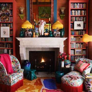 A-List 2024 - ELLE DECOR very cozy closeup of a sitting area with red plank walls and a white marble fireplace with a large blue and red mirror over it and jade colored objects and elephant lamps with matching yellow fringe shades and in front of the fire is a small colorful chair and matching chaise with red fringe all on a multicolored carpet and two tall bookcases on either side of the fireplace also painted red