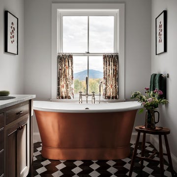 white bathroom with window with a mountain view, half curtains, deep copper tub, side table with vase and flowers, wood cabinet with marble top and sink, black and white diamond floor tiles, pendant over tub