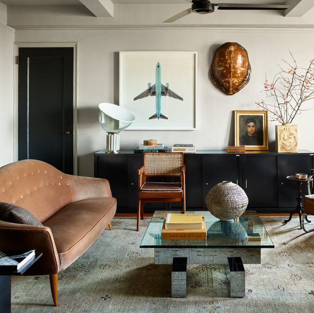 A-List 2024 - ELLE DECOR a living room with a long black console on the back wall with art and book around it and a worn leather sofa on the left and a low geometric cocktail table with a glass top in fron on a faded rug and old metal and leather armchair at right