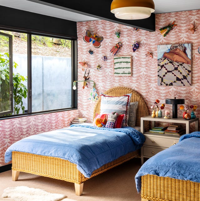 A-List 2024 - ELLE DECOR twin rattan beds with oval headboard and blue coverlets against a pink patterned wallpaper and a nightstand in between and a long window and bug artwork and objects on the wall