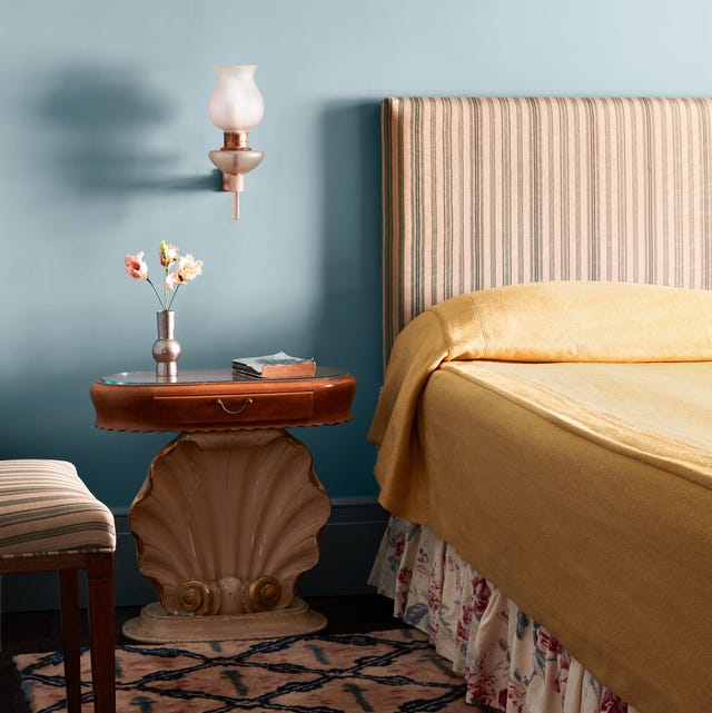 A-List 2024 - ELLE DECOR bedroom with blue wall and a bed with tall striped headboard and flowered bed skirt and plain white coverlet and a patterned pink and blue carpet and a nightstand with an oval wood top and a seashell pedestal
