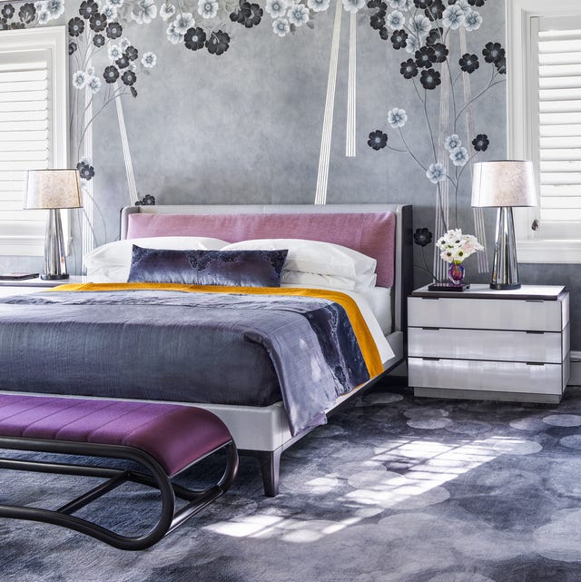 A-List 2024 - ELLE DECOR bedroom in soft slate blue and lilac colors with a mottled slate blue carpet and a similar darker colored bedcover and a pink velvety insert into the headboard and a dark purple bench with black bottom at the foot and a white three drawer nightstand and the wall has a wallcovering in gray with black and white simple flowers on parts of it
