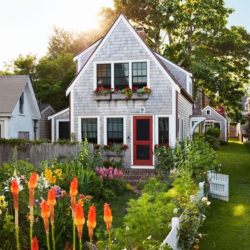 David Cafiero - ELLE DECOR the exterior of a cedar shingled two story cottage with window boxes with red flowers, a red screen door, and brick steps, a white picket fence with roses surrounds tall orange flowers, yellow day lilies, pink foxglove
