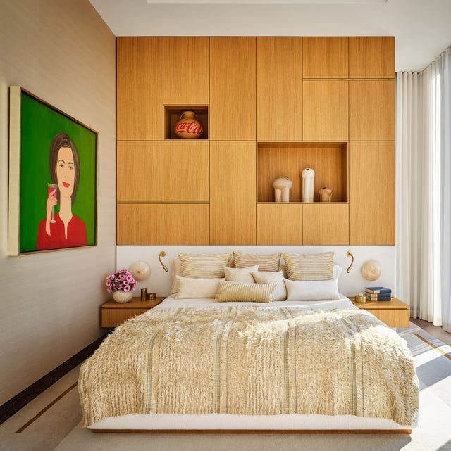 A-List 2024 - ELLE DECOR an airy high ceilinged bedroom with a bed and light wood paneled wall with some inset shelves and cabinetry and on the right is a large painting with a green background of a woman in red holding a glass of wine