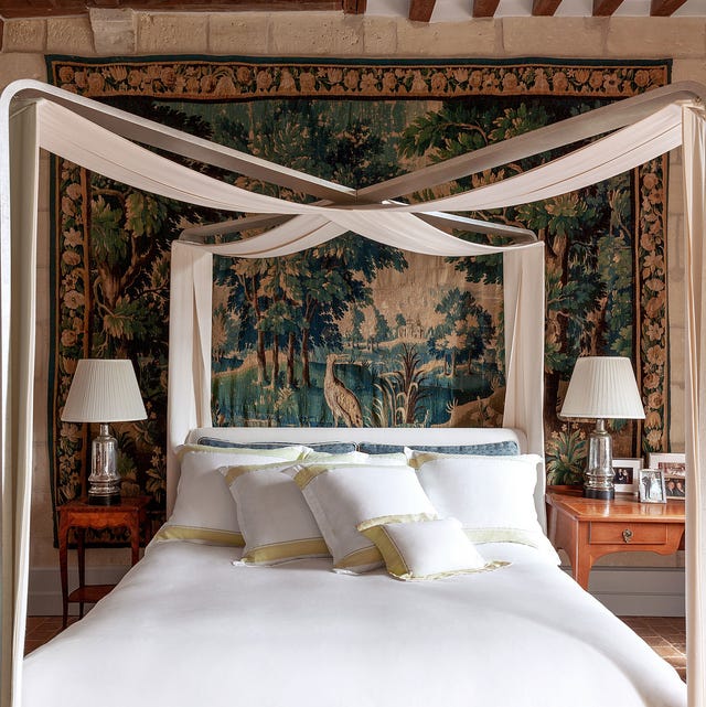 A-List 2024 - ELLE DECOR a white linen bed with a ribbon canopy draped in an x overhead and a large tapestry on the wall behind and two small mismatched sidetables and silver base lamps