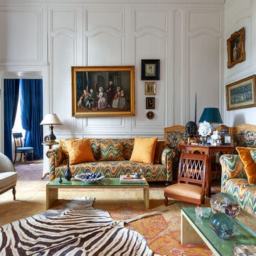 Robert Couturier ELLE DECOR a great room with numerous framed artworks on the paneled walls, two sofas with a swirly colorful print, a wooden chair and an armchair, green glass topped cocktail tables, zebra print rug, deep blue curtains