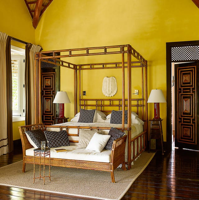 A-List 2024 - ELLE DECOR yellow painted room with bamboo cane four poster bed and matching empire looking settee at the foot and dark wood floors and carved black doors with wood insets