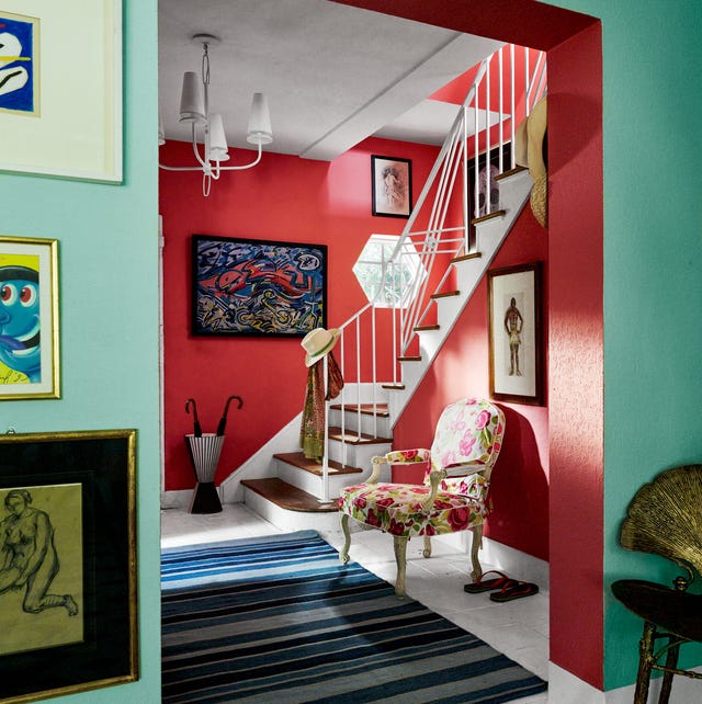 A-List 2024 - ELLE DECOR view from a green painted room with artwork into a red painted entry with stairs going up and a floral fauteuil next to it and a striped rug in blue hues and artwork on the walls and a simple chandelier