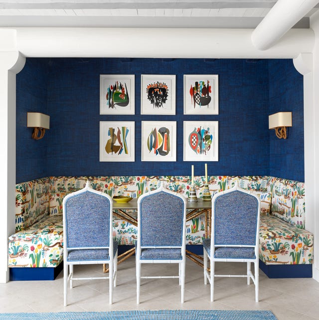 A-List 2024 - ELLE DECOR a happy flower printed banquette that wraps around three dark blue walls with six white framed artworks in a grid and tall table at center and three blue chambrey like upholstered chairs pulled up to it and