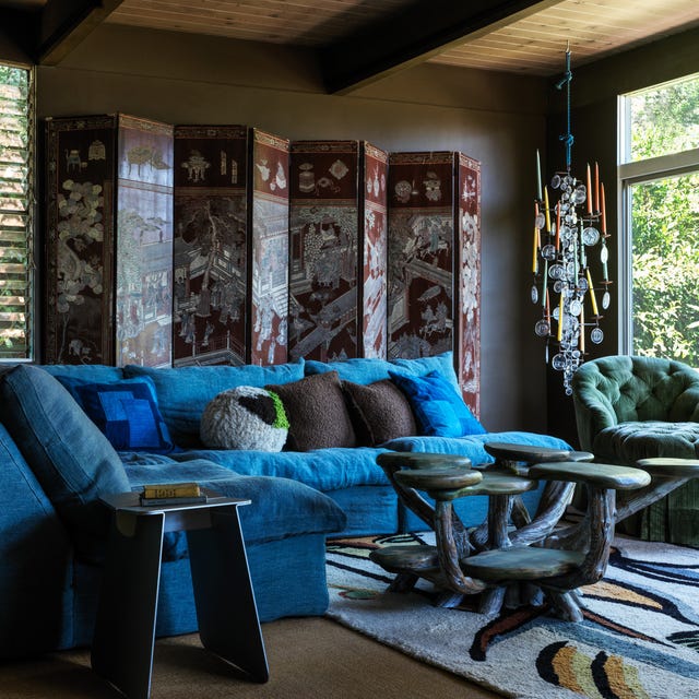 A-List 2024 - ELLE DECOR moody living space with bright blue velvety small sectional sofa and a cocktail table with several arms with tops and a large screen against the wall behind the sofa