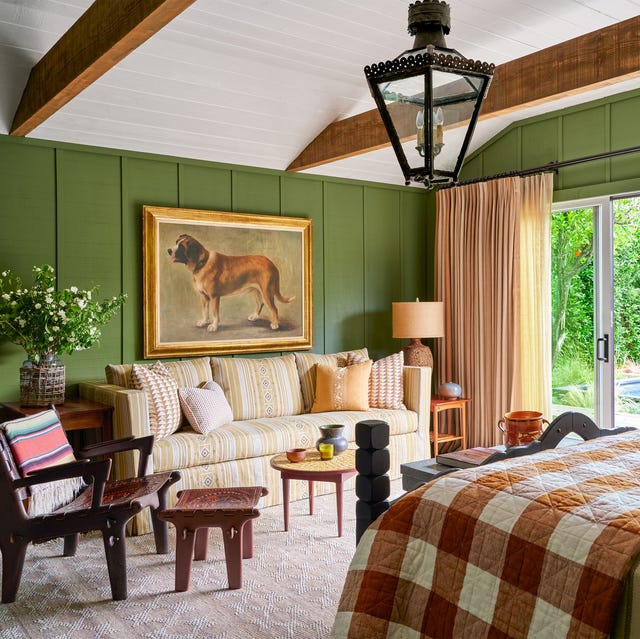 A-List 2024 - ELLE DECOR a bedroom with green paint walls with a large photo of a dog on one side and a light striped sofa with low wood tables in front and then opposite a bed with a brown and white large check blanket at the foot