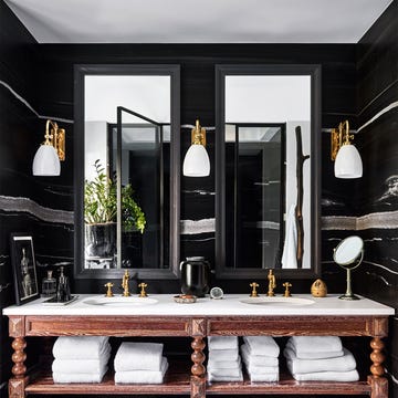 a bathroom with black walls, three sconces flanking two mirrors, wooden vanity with turned legs and two shelves holding towels and baskets, a white marble top with two small round sinks with brass fittings