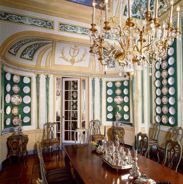 A-List 2024 - ELLE DECOR a large ornate room with doomed ceilngs and a chandelier and and polished mahogany table with silver service on top and chairs around with striped seats and on the walls are green panels hung with rows of plates