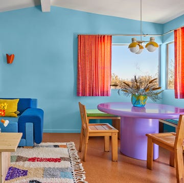 colorful living room with light blue walls and a dark blue sofa and a purple pedestal table and wood chairs and burnt orange curtains on half size window and a colorful rug under the cocktail table