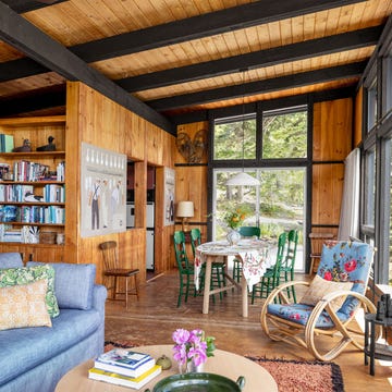 a living and dining area has a blue sofa with accent pillows, a bentwood rocker with a flowered cushion, wood paneled walls and beamed ceiling, a dining table with green painted chairs, a built in book shelf