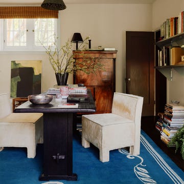 a home office with a dark wood desk and two white square shaped armless chairs, open shelving with books on one wall, closed secretary desk, blue patterned rug