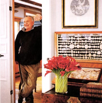 older man in a black sweater and baggy corduroys leans against a doorway with a small table in the foreground with a vase of red flowers and some old books on it