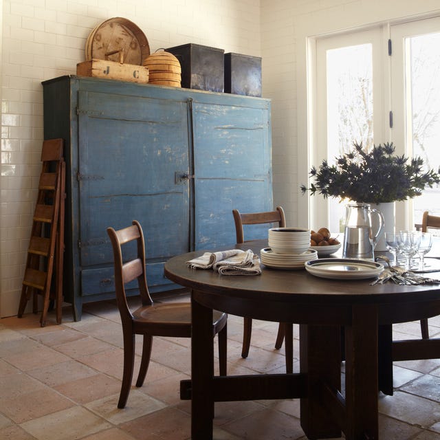A-List 2024 - ELLE DECOR a round wood table with plates and glasses on it and a rustic blue painted chest behind it with old fashioned boxes and bins and french doors to the back right