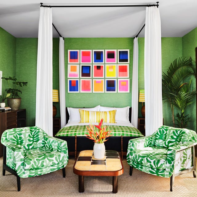 A-list 2024 - ELLE DECOR a bedroom with lots of green including a canopy bed with white sheers and a green check blanket and two palm tree patterned green and white armchairs at the foot and a sisal carpet on wood floors and a colorful array of frames over the bed