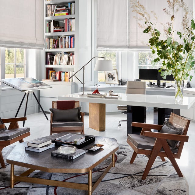 A-List 2024 - ELLE DECOR study or den with long white shiny lacquer tri pedestal table with a midcentury low cocktail table and chair over a geometric black and white rug and an easel with flat open book at back and a white inset bookshelf