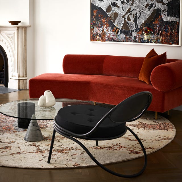 A-List 2024 - ELLE DECOR swishy reddish orange velvet armless sofa in a living area with a round rug that looks like a tortilla and a black wire chair and glass cocktail table with three different shaped legs and a large artwork in complementary colors on the wall