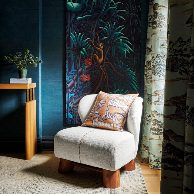 A-List 2024 - ELLE DECOR white upholstered chair with thick wood beam legs on a light striated carpet agsinst a blue textured wall with a monkey artrwork and toile curtains in a moody corner