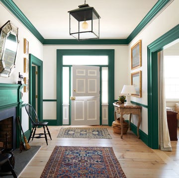an entry hall has off white walls with rich green moldings and woodwork, a dutch door painted light gray, a fireplace, two black wooden chair, two caucasian rugs, a marble topped half moon console, and a gallery lantern