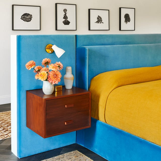 A-List 2024 - ELLE DECOR upholstered platform bed in a blue velvet with a yellow cover and a floating mounted two drawer night stand with a small white vase with flowers on it and a small mounted white spotlight
