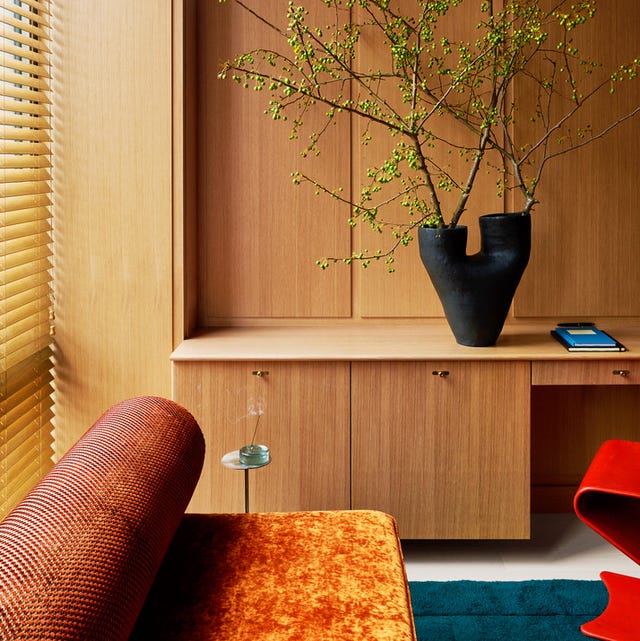 A-List 2024 - ELLE DECOR details of pale honey colored built in cabinetry with a black vase and branches on it and a bed in the foreground with an orange and blue modern bed with roll head and a red lacquer potato chip type chair peeking out from the right