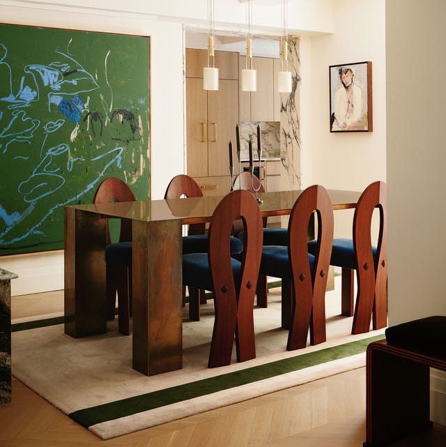 A-List 2024 - ELLE DECOR a high glossy wood dining table with chair pulled up to it with ankh shapped backs and blue seats on a cream rug with two green stripes and a large green splatter painting on the wall and glass cabinetry