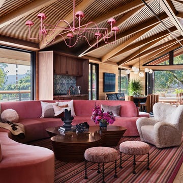 living room with slanted, beamed wooden ceiling, curved pink sectional sofa, curved cocktail table, boucle chair, two stools, striped rug, pink chandelier, large windows, dining table and kitchen island in background