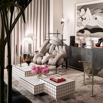 living room with cocktail table formed of three cubes of small white squares, chain mail chair with metal base, chair with interweaving taupe fabric tubes, black sideboard with large artwork above, end table with lamp