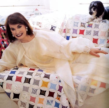 a smiling person lying on a bed with a little dog looking over from the top of the sofa