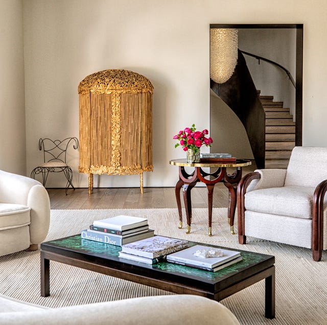 A-List 2024 - ELLE DECOR a living room with mismatched white armchairs and a swoopy curvy white sofa and rectangular cocktail table on a white striated rug and a gold encrusted rounded top console against the back wall where there is a doorway leading to a swoopy staircase upstairs