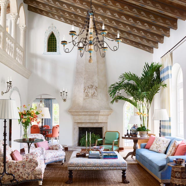 A-List 2024 - ELLE DECOR very dramatic high ceilinged living room with ornate wave designed beams up top and a tall fireplace hood and a wide upholstered cocktable table at center with a blue sofa on one side and two matching floral patterned armchairs to the left and a tall plume of palm tree at back and very long white and blue curtains next to arched windows