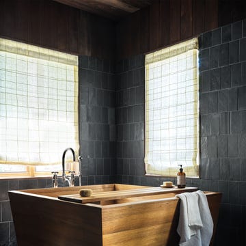 a bathroom has black limestone tiles on the floor and walls, two windows with linen roman shades in a small square pattern, a deep square cedar tub with a wooden tray holding soap and a dispenser, chrome fittings