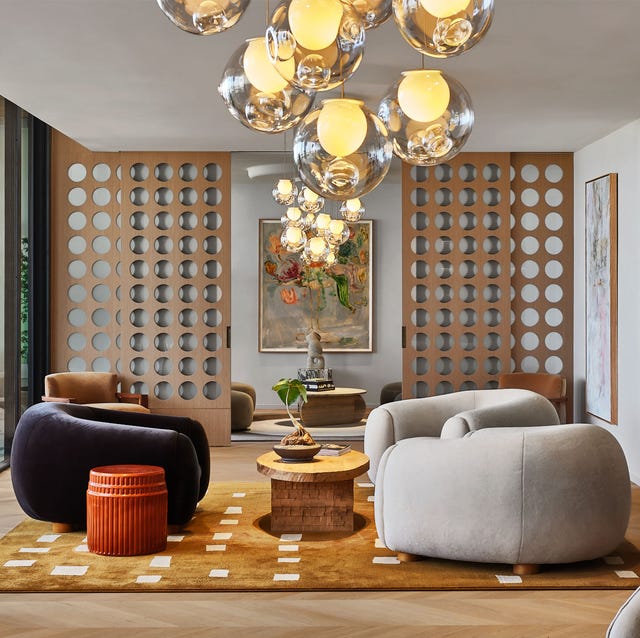 A-List 2024 - ELLE DECOR a living room with a lattice screen behind and large bean bag like armchairs on a caramel colored rug with white squares and a bubble chandelier