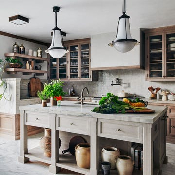 kitchen with an open door to a terrace, large marble topped island with fluted edge, sink, vases stored underneath, two pendants, long countertop with wood cupboards below and glass cabinets above